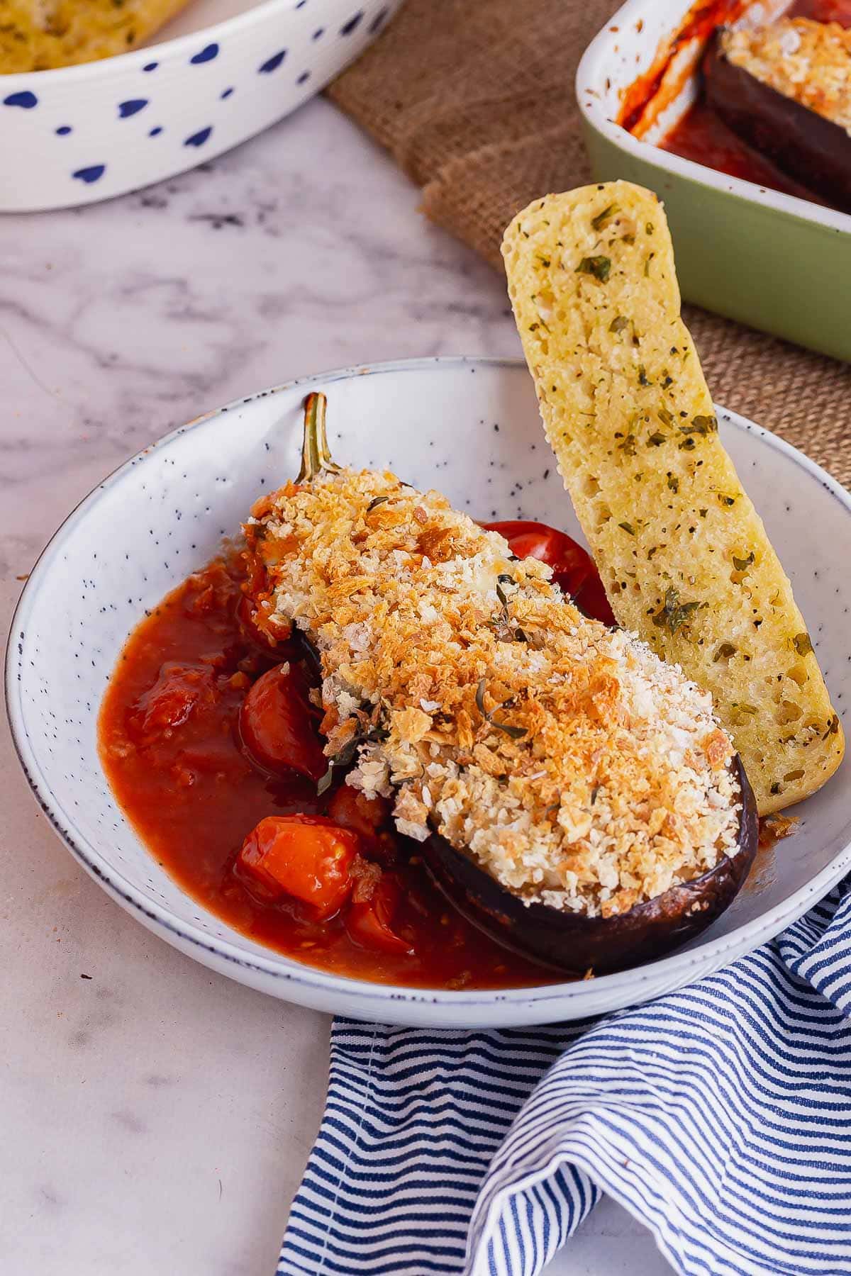 Blue bowl of cheesy aubergine with tomato sauce and garlic bread on a marble background