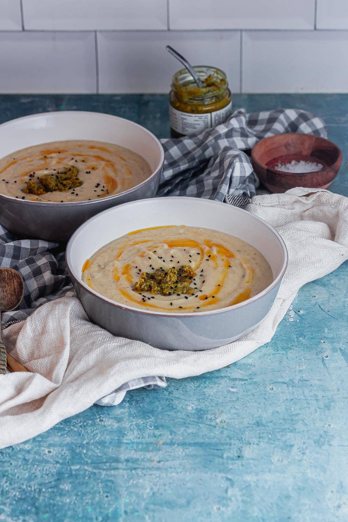 Two bowls of cauliflower soup on a blue surface in front of a white brick tile wall