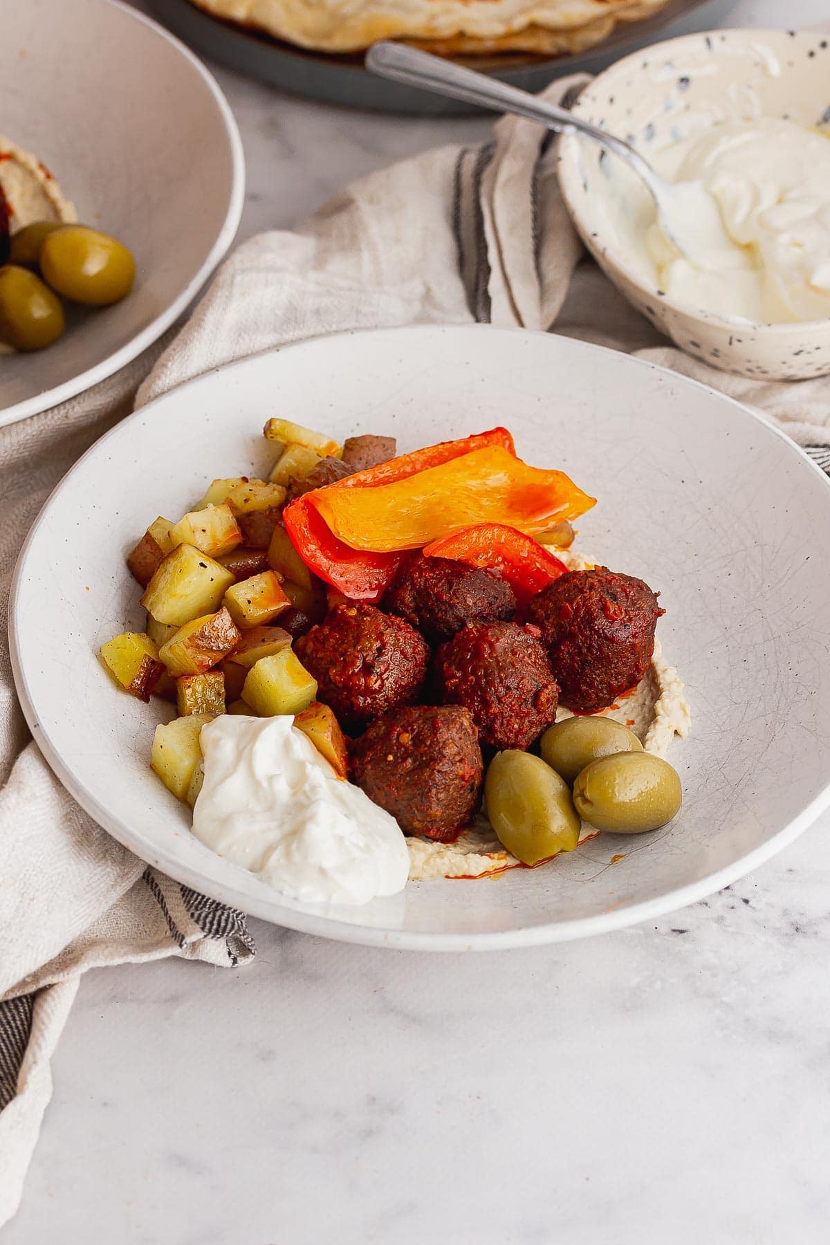 White bowl of spicy meatballs with potatoes and hummus on a marble background